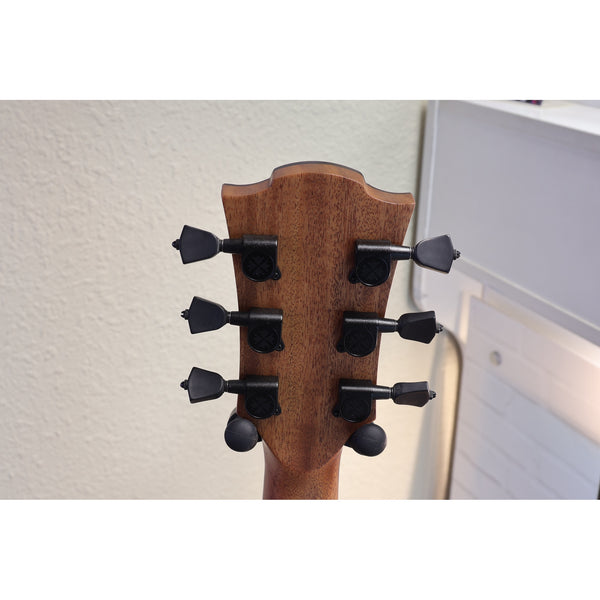 Close-up of the headstock of a wooden stringed instrument, likely a guitar or ukulele, showing six black tuning pegs against a light wall background.