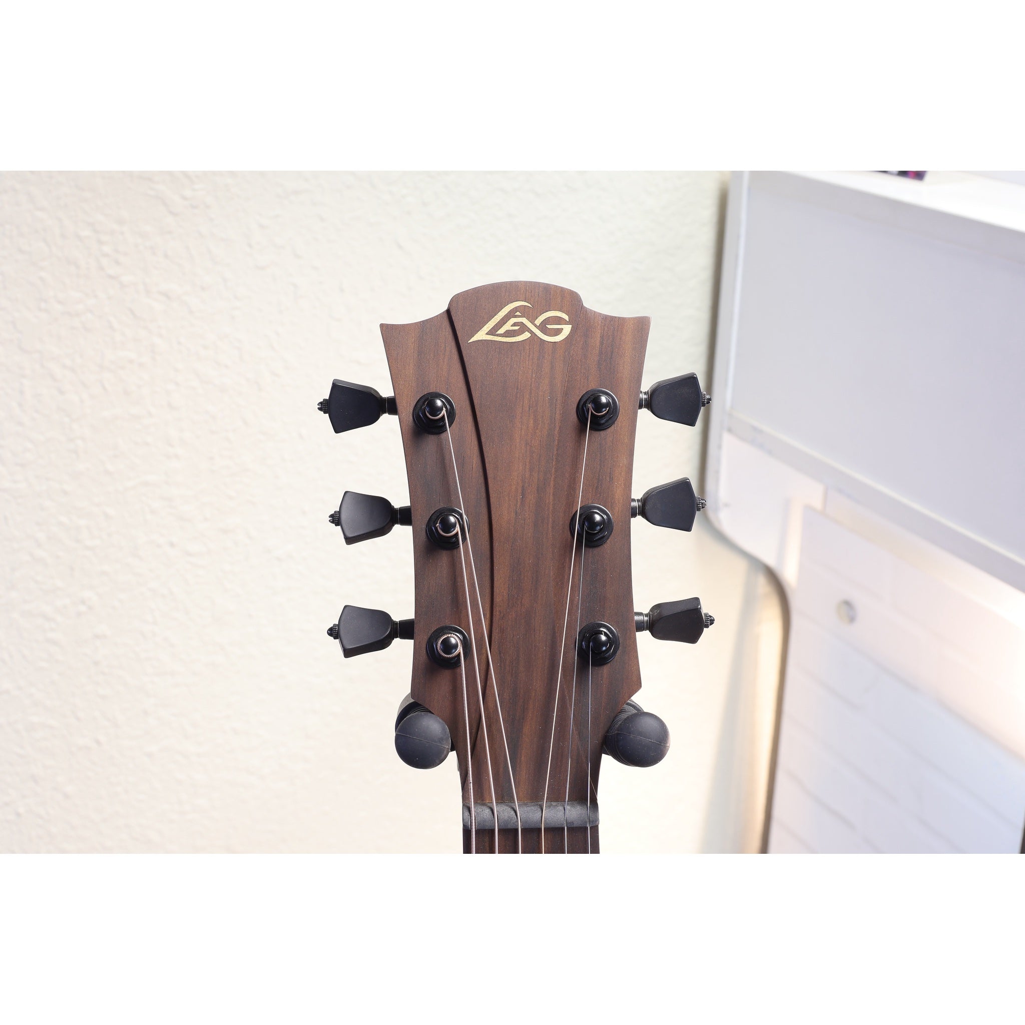 Close-up of the headstock of an LG acoustic guitar, showing the dark brown wood grain, six black tuning pegs, and the gold LG logo, set against a light-colored wall.
