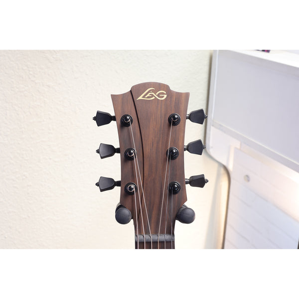 Close-up of the headstock of an LG acoustic guitar, showing the dark brown wood grain, six black tuning pegs, and the gold LG logo, set against a light-colored wall.